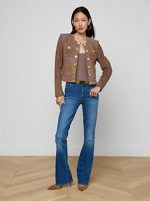 A woman with straight dark hair and a neutral expression stands indoors against a plain wall, wearing the Jayde Collarless Jacket over a taupe top, blue flared jeans, brown pointed shoes, and large hoop earrings.