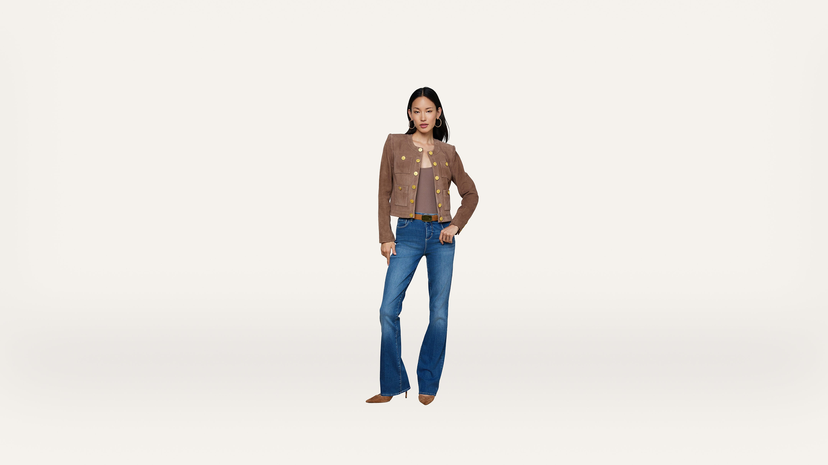 A woman stands against a plain white background, wearing a brown jacket, a light top, blue flared jeans, and brown shoes, with one hand on her hip.