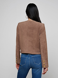 A woman with long dark hair, wearing the Jayde Collarless Jacket—a brown suede jacket with gold buttons on the sleeves—paired with blue jeans, stands facing away against a light background. This stylish women’s layering piece is collarless for a modern look.