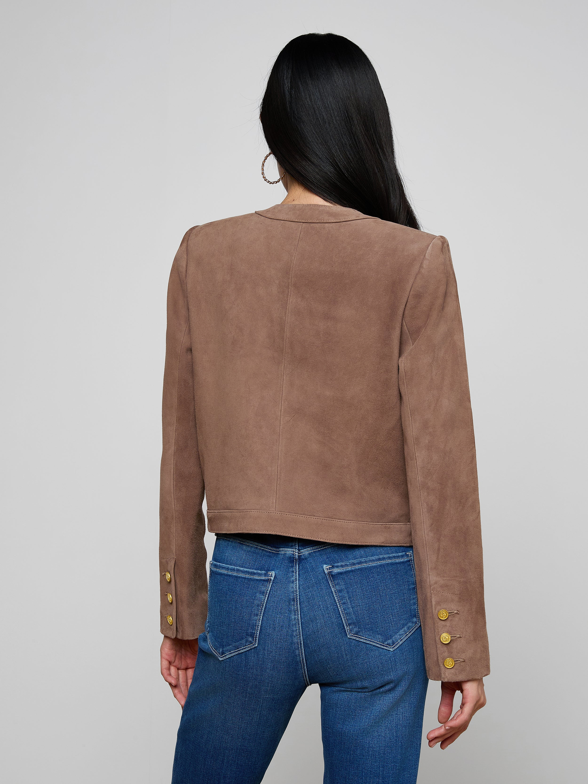 A woman with long dark hair, wearing the Jayde Collarless Jacket—a brown suede jacket with gold buttons on the sleeves—paired with blue jeans, stands facing away against a light background. This stylish women’s layering piece is collarless for a modern look.