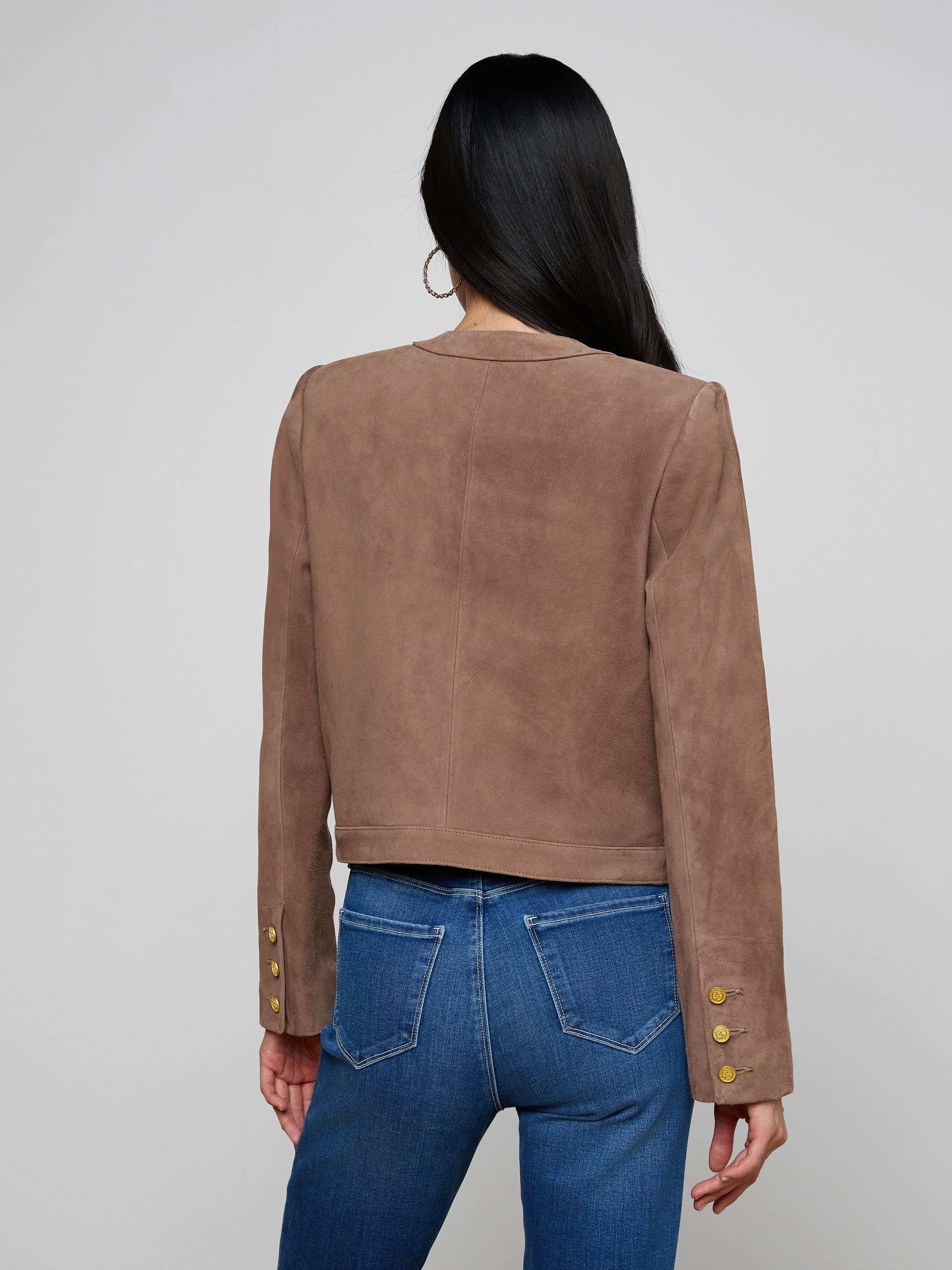 A woman with long dark hair, wearing the Jayde Collarless Jacket—a brown suede jacket with gold buttons on the sleeves—paired with blue jeans, stands facing away against a light background. This stylish women’s layering piece is collarless for a modern look.