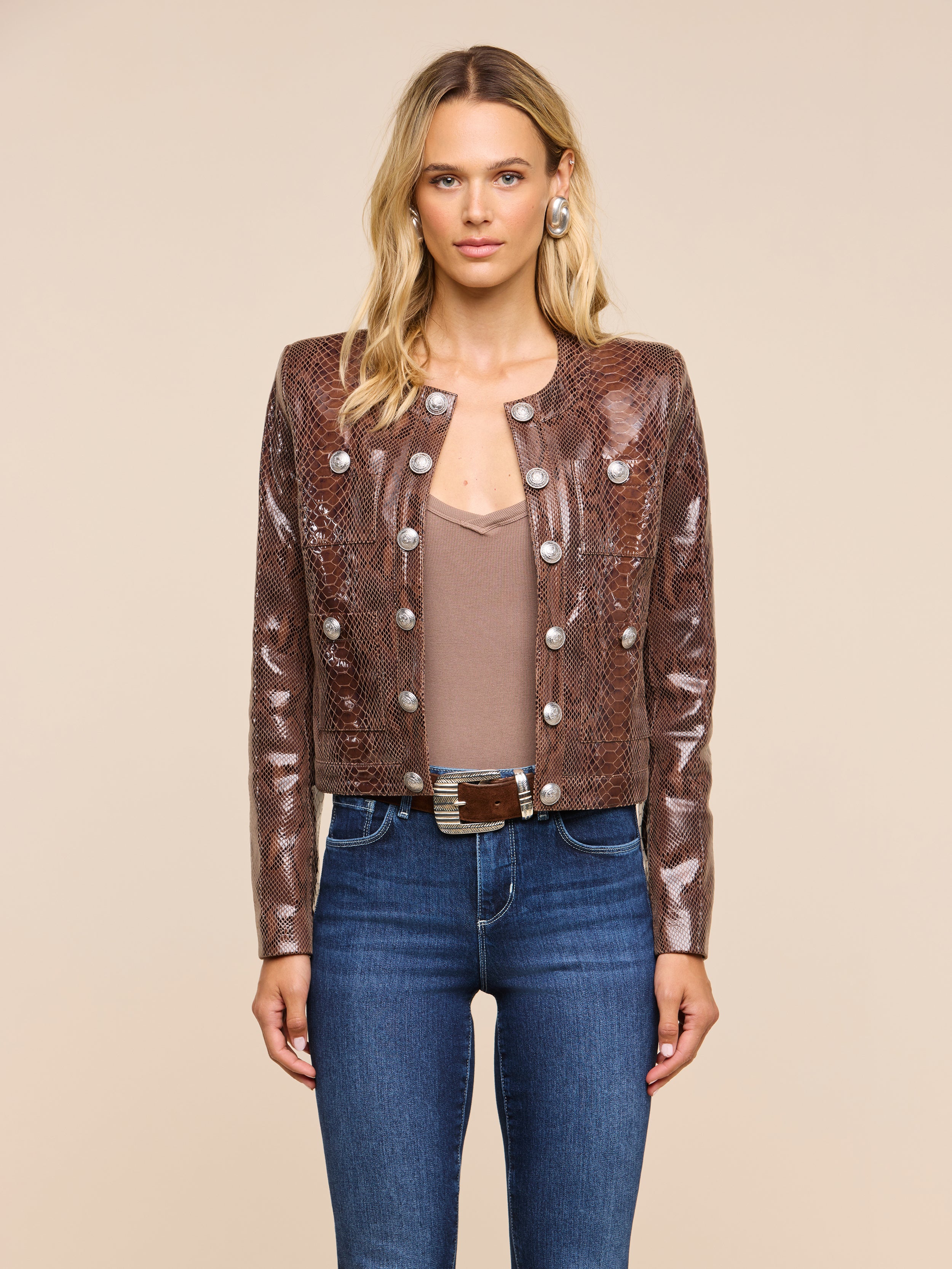 A woman stands facing forward, wearing the Jayde Coated Suede Jacket—a textured brown snake print piece with metallic buttons—paired with a beige top, blue jeans, and a belt. She has blonde hair and a neutral expression, posed against a plain light background.