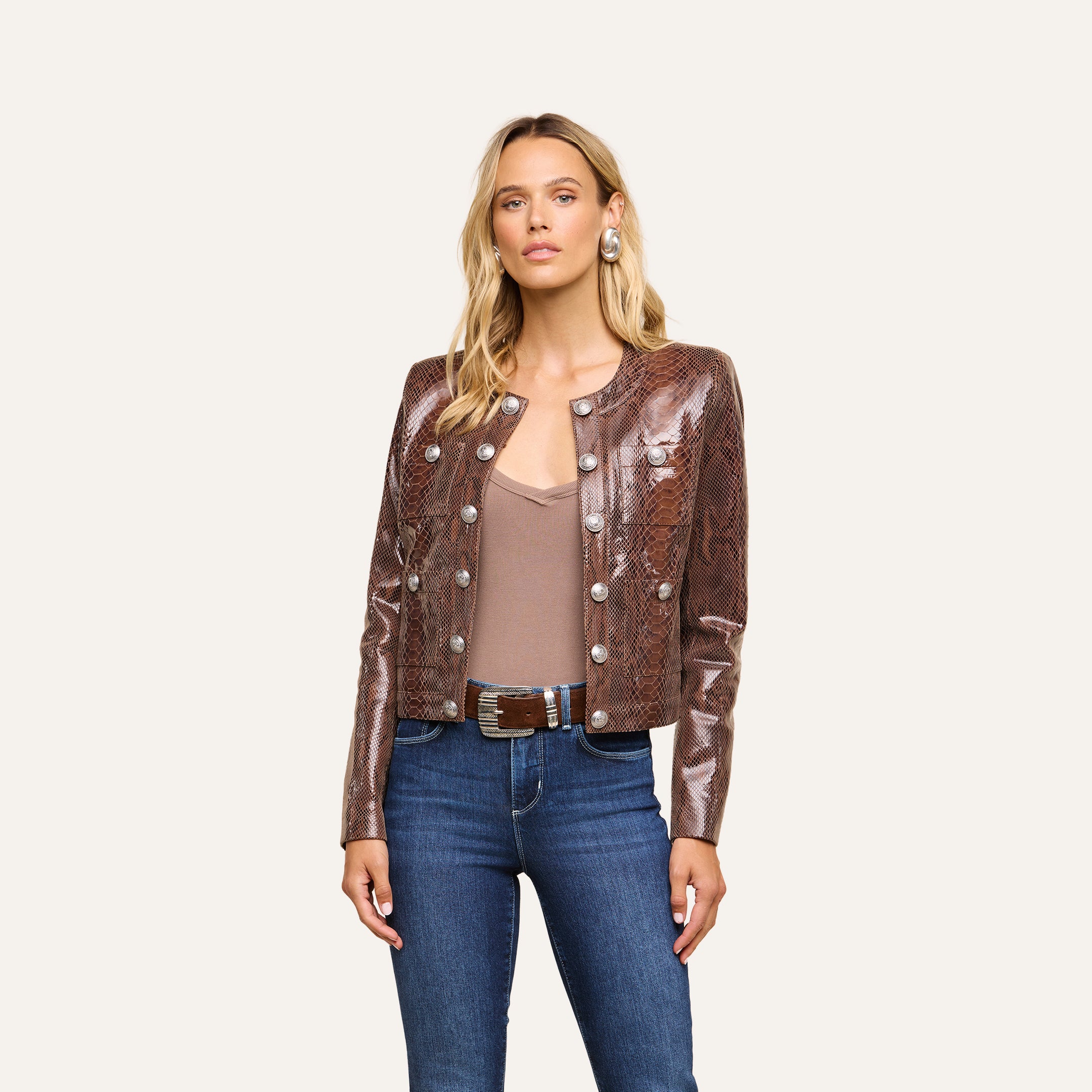 A woman wearing a shiny brown Jayde Coated Suede Jacket with silver buttons, a beige top, blue jeans, and large round earrings stands against a plain light background.