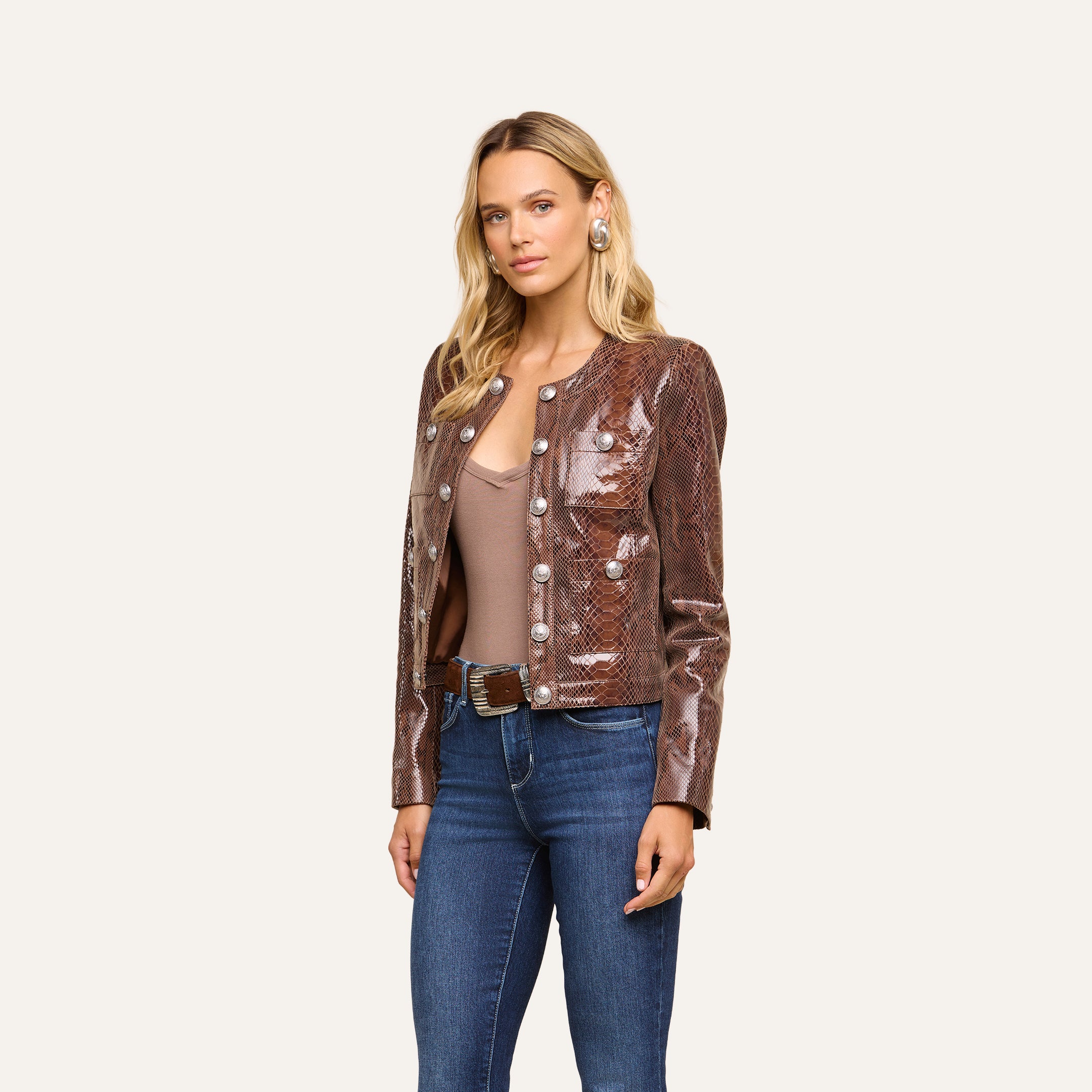 A woman with long blonde hair wears the Jayde Coated Suede Jacket, paired with a taupe top, blue jeans, and a brown belt, as she stands against a plain light background.