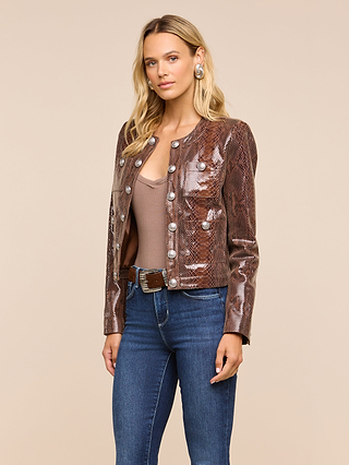 A woman wearing the Jayde Leather Jacket—a shiny brown snake print piece with a high-shine finish—paired with a taupe top, blue jeans, and large silver earrings stands against a plain beige background.