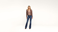A woman stands against a plain white background, wearing a brown patterned jacket, a taupe top, blue flared jeans, and brown shoes. She has straight blonde hair and a neutral expression.