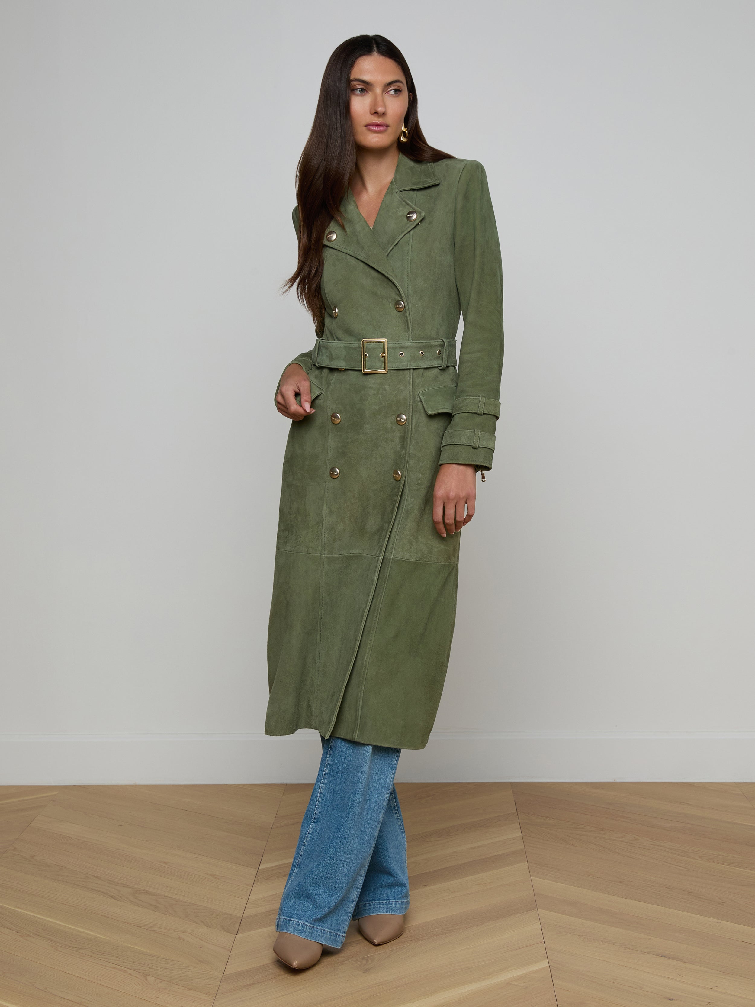 A woman models the Celina Suede Trench Coat in olive green, styled over blue jeans and beige shoes, standing against a plain white background.
