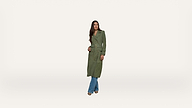 A woman wearing a long green trench coat over blue jeans and brown shoes stands against a plain white background.