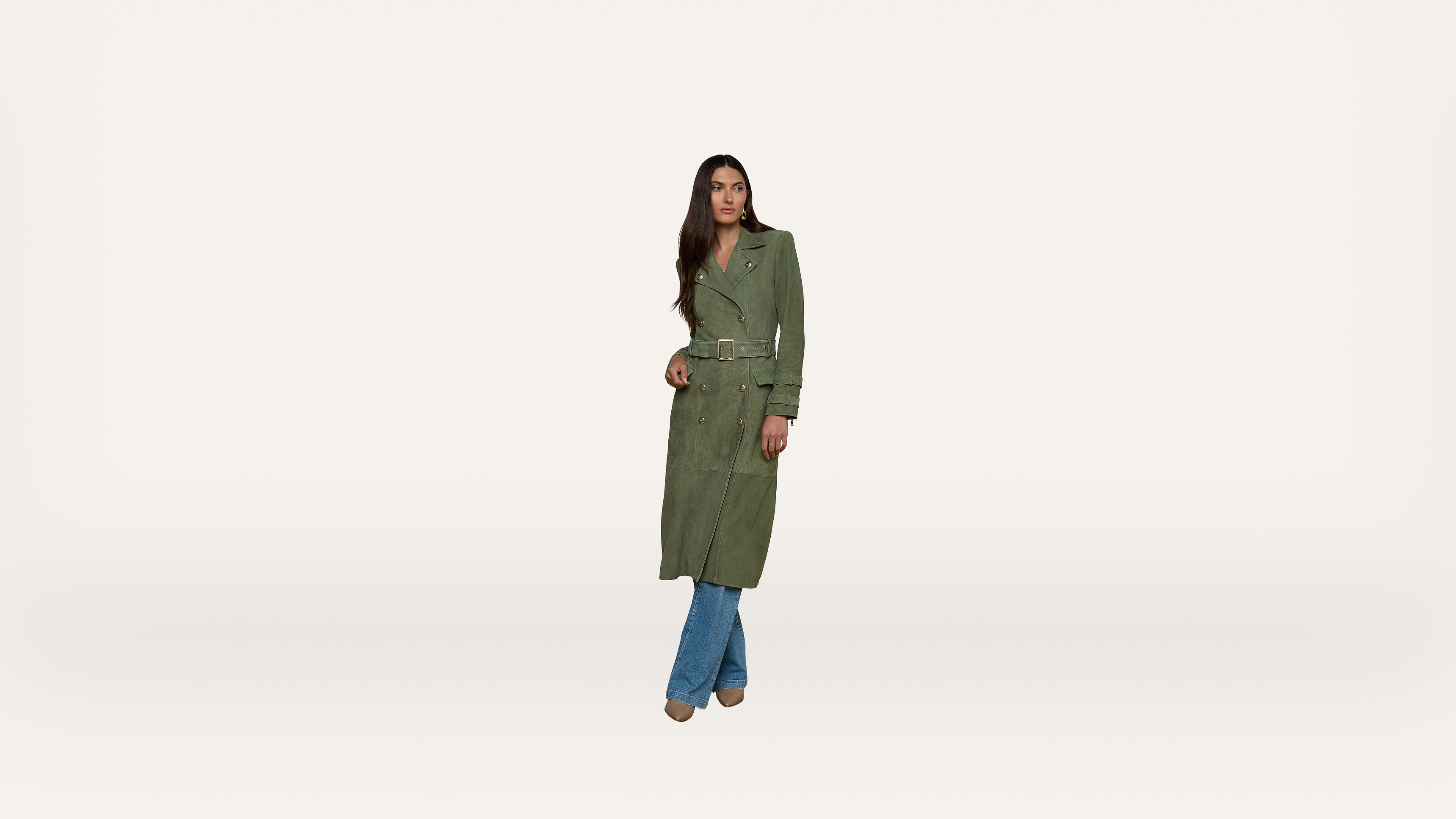 A woman wearing a long green trench coat over blue jeans and brown shoes stands against a plain white background.