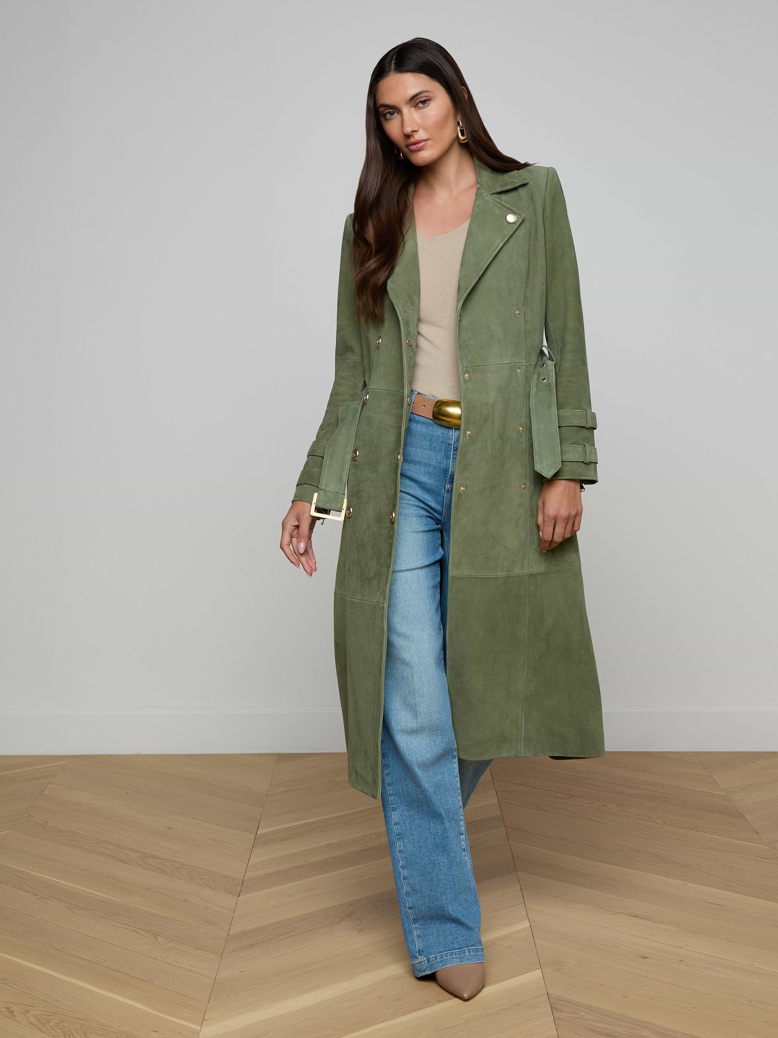 A woman wearing a long olive green Celina Suede Trench Coat, beige top, blue flare jeans, and tan shoes stands against a plain light background.