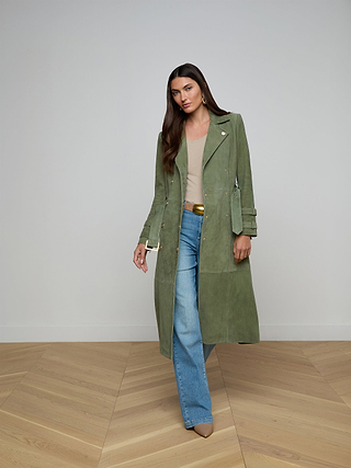 تقف امرأة داخل المنزل مرتدية بنطال جينز «Petite Scottie Coated Jean» مع معطف ترنش طويل أخضر، وقميص بيج، وحزام بني فاتح، وحذاء بكعب بني. ولديها شعر بني طويل، وأقراط حلقات ذهبية، وتقف أمام جدار سادة فاتح اللون وأرضية خشبية.