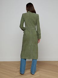 A woman stands facing away, wearing a long green Celina Suede Trench Coat belted over blue jeans. She is posed against a plain white background, highlighting this timeless wardrobe essential.