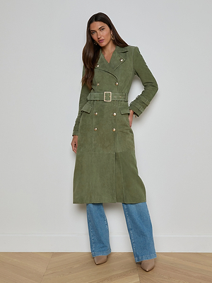 A woman wears the Celina Suede Trench Coat in olive green with gold buttons and a belt, styled with blue jeans and beige heels against a white wall—a timeless look for any wardrobe.
