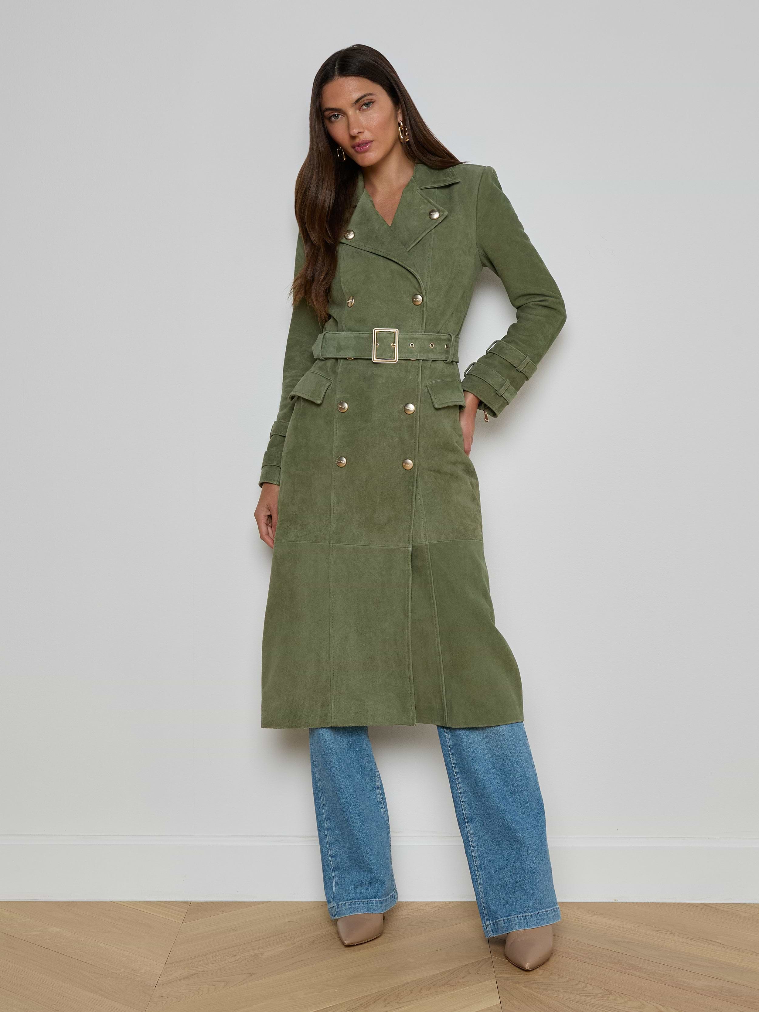 Woman wearing a long olive green Celina Suede Trench Coat over blue jeans and tan heeled shoes, standing against a plain white background—a perfect addition to any timeless wardrobe.