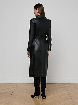 A woman stands indoors with her back to the camera, wearing the L'AGENCE Celina Leather Trench Coat and black heeled boots. The scene features light wood flooring and a plain white wall.