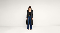 A woman wearing sunglasses, a long black coat, black top, blue wide-leg jeans, and black heels stands against a plain white background.
