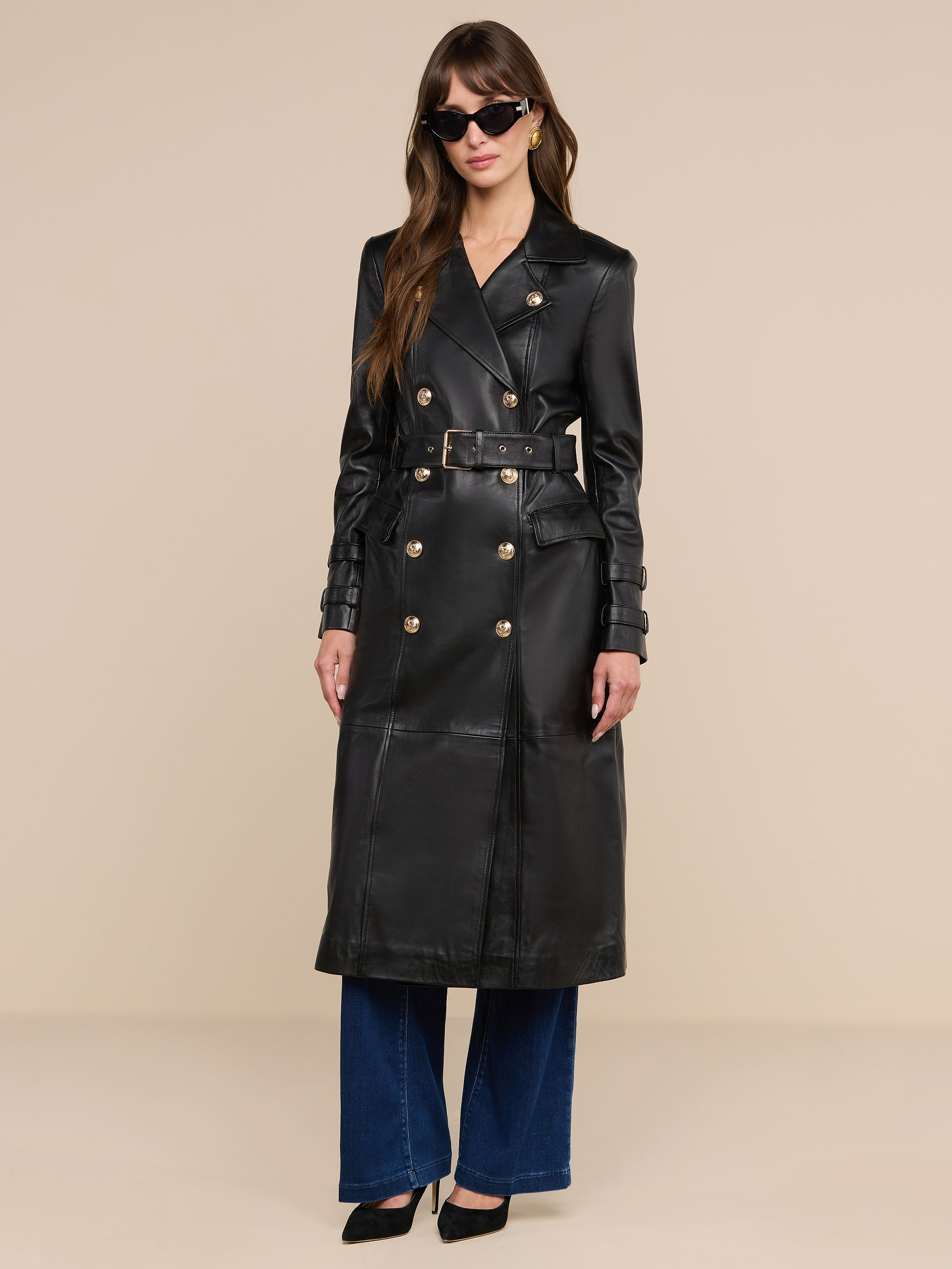 Una donna è in posa su uno sfondo beige e indossa il trench in pelle Celina nero con bottoni dorati, abbinato a jeans blu a gamba larga, tacchi neri e occhiali da sole.