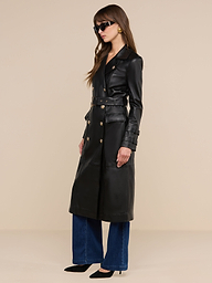 Una donna è in posa su uno sfondo beige a tinta unita e indossa il trench in pelle Celina, caratterizzato da bottoni dorati, abbinato a jeans blu a gamba larga, tacchi neri, occhiali da sole e grandi orecchini rotondi.
