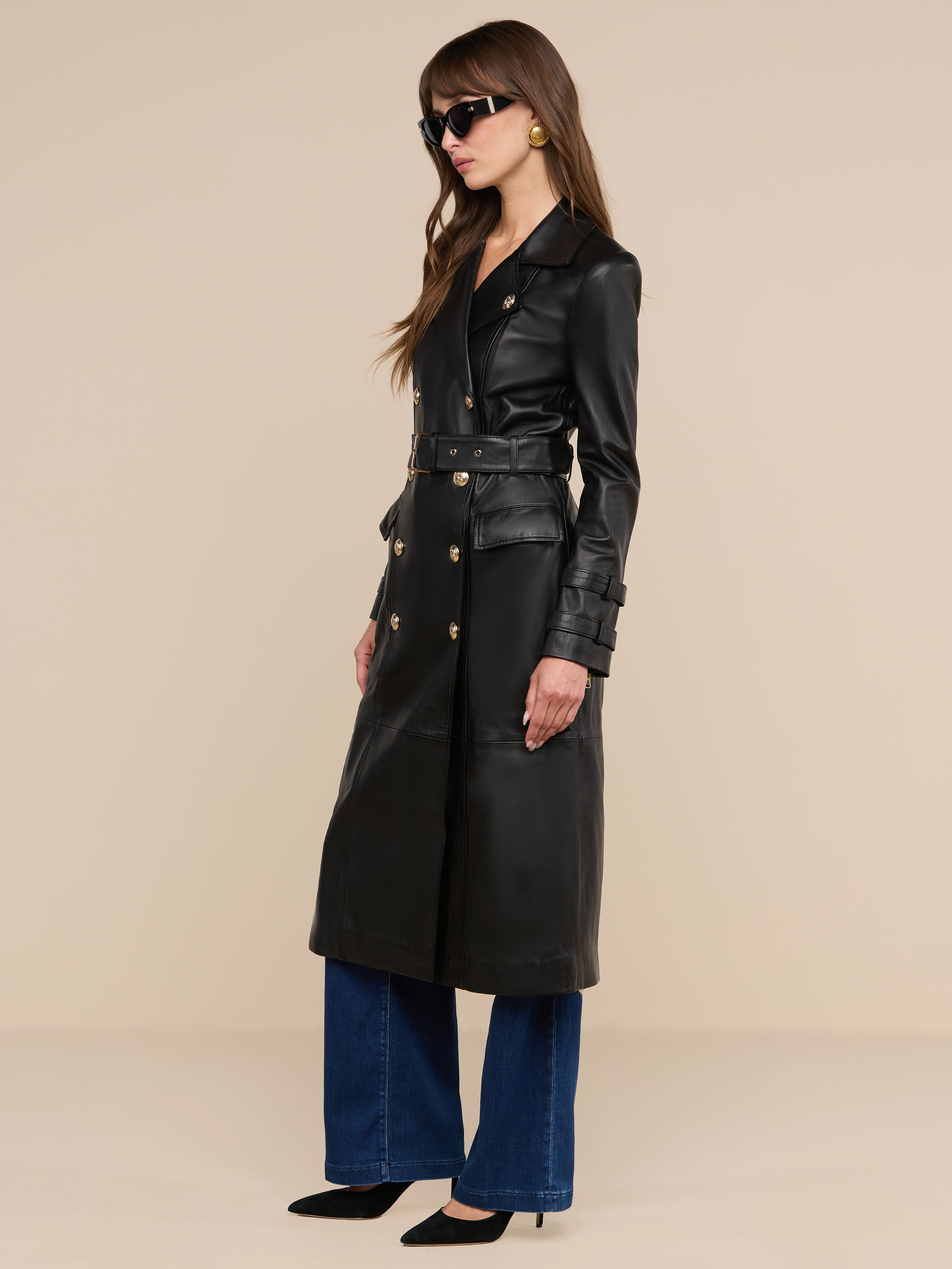 Una donna è in posa su uno sfondo beige a tinta unita e indossa il trench in pelle Celina, caratterizzato da bottoni dorati, abbinato a jeans blu a gamba larga, tacchi neri, occhiali da sole e grandi orecchini rotondi.