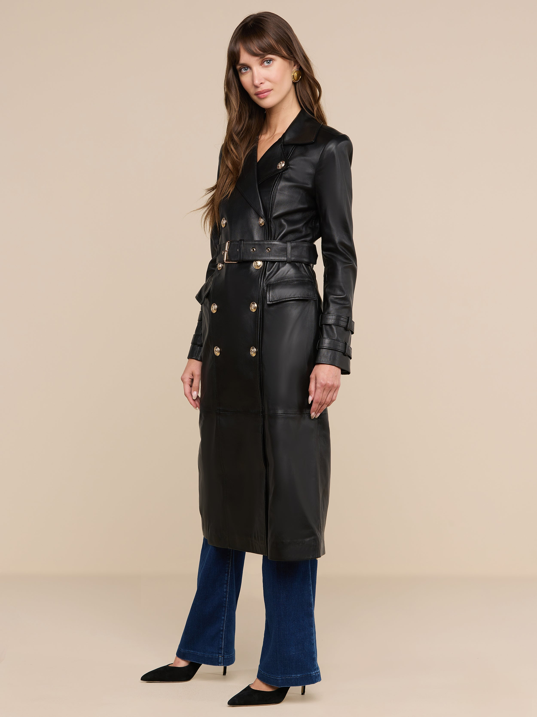 Una donna indossa il trench in pelle Celina, un modello di alta qualità di colore nero con bottoni dorati, abbinato a jeans blu e tacchi neri, in piedi davanti a uno sfondo beige a tinta unita.