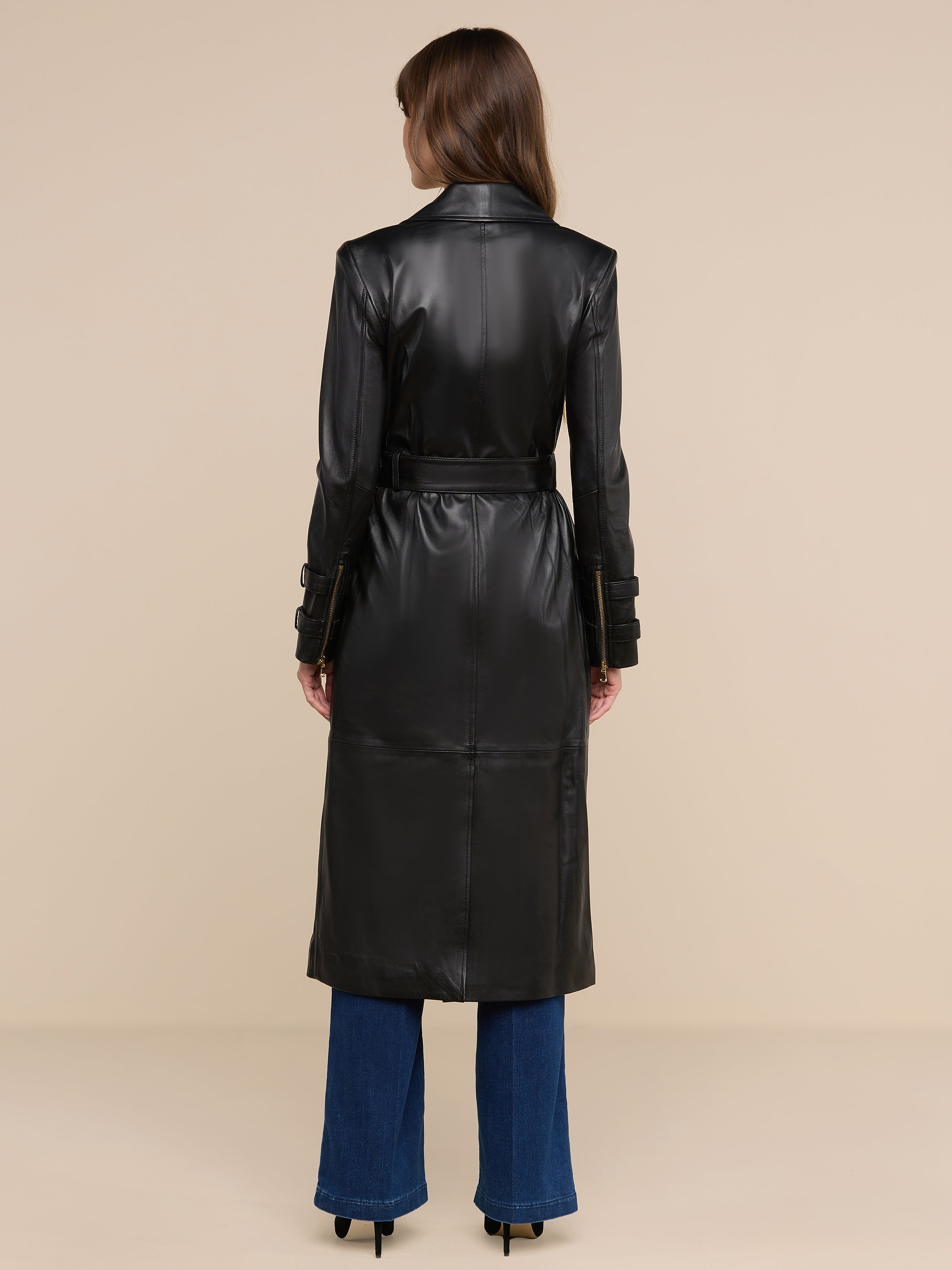 Una donna è di spalle, indossa il trench in pelle Celina nero con cintura in vita, abbinato a jeans blu scuro e tacchi neri, su uno sfondo beige a tinta unita.