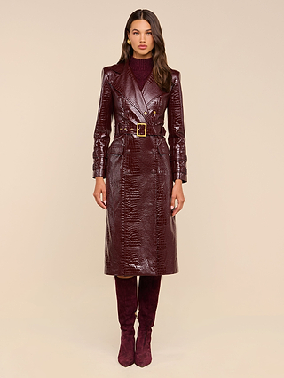 Woman wearing the Celina Trench Coat in shiny burgundy vegan leather with crocodile embossing and a matching belt, styled with a maroon turtleneck and burgundy knee-high boots, standing against a neutral background—a striking look that perfectly captures Fall 2026 fashion.