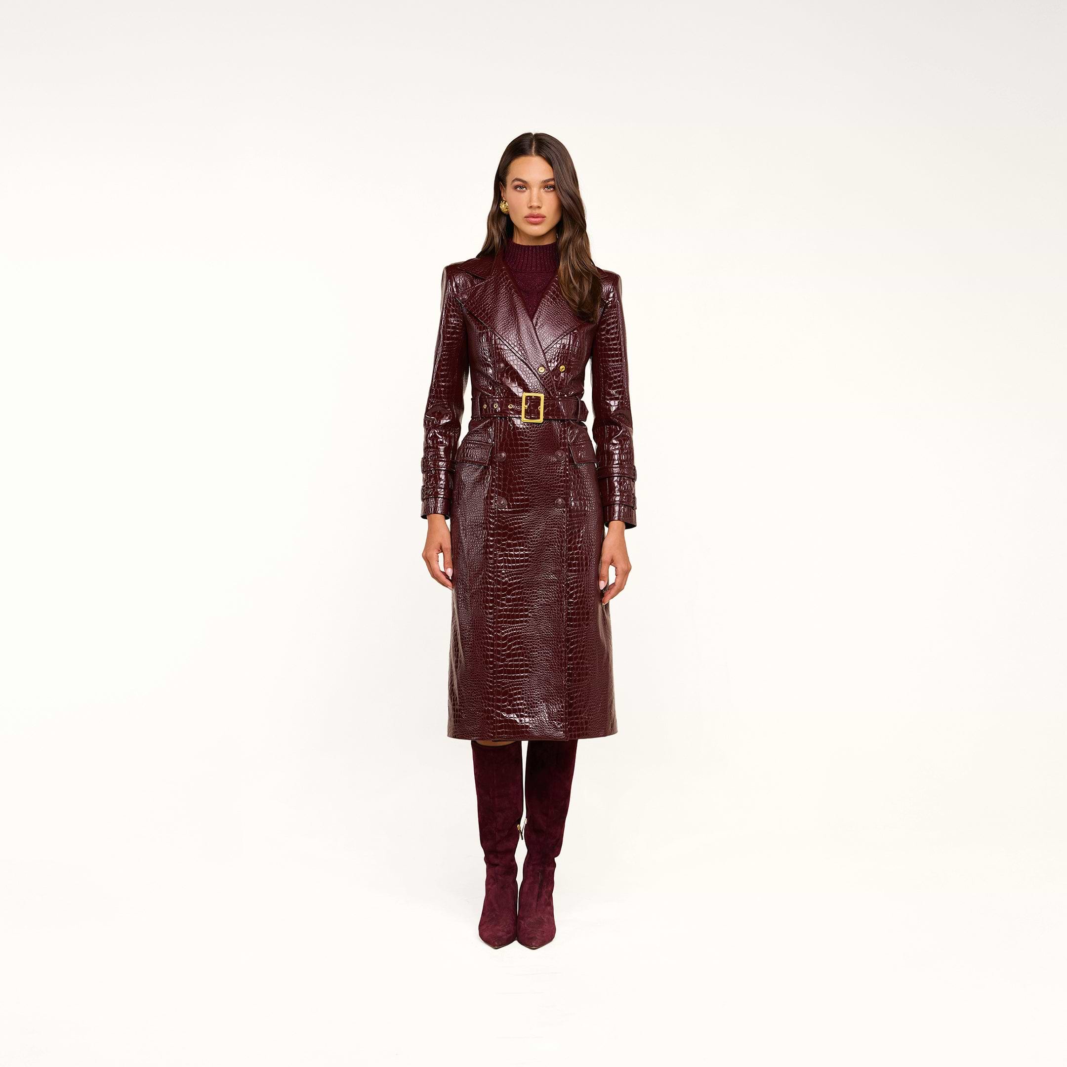 Woman wearing the Celina Trench Coat in shiny burgundy vegan leather with crocodile embossing and a matching belt, styled with a maroon turtleneck and burgundy knee-high boots, standing against a neutral background—a striking look that perfectly captures Fall 2026 fashion.
