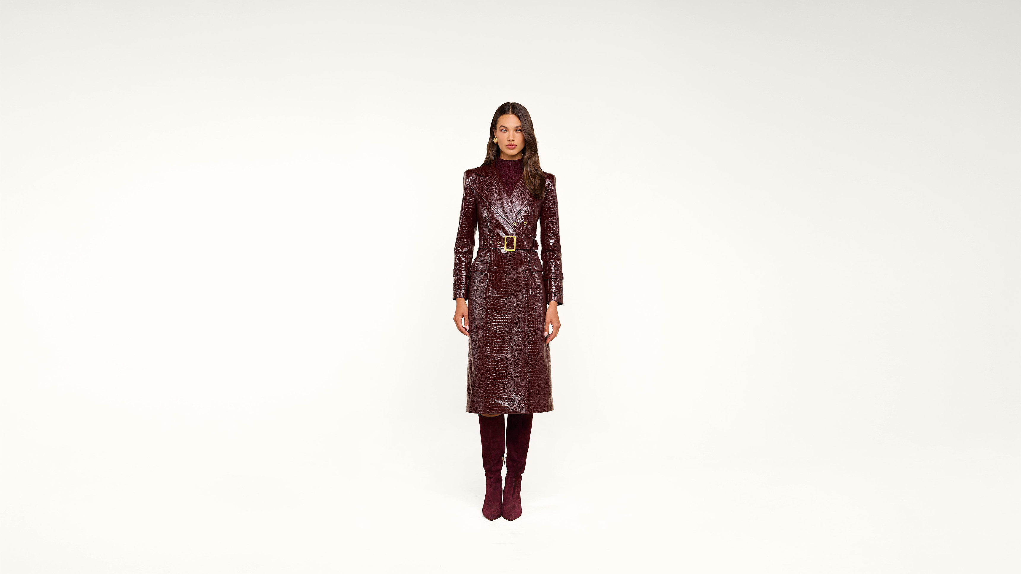 Woman stands facing forward, wearing a shiny, belted burgundy trench coat and matching knee-high boots against a plain white background.