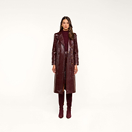 A woman stands facing forward, wearing the Celina Trench Coat—a shiny burgundy vegan leather piece with crocodile embossing—layered over a matching dress and knee-high suede boots, set against a plain beige background for a striking look inspired by Fall 2026 fashion trends.