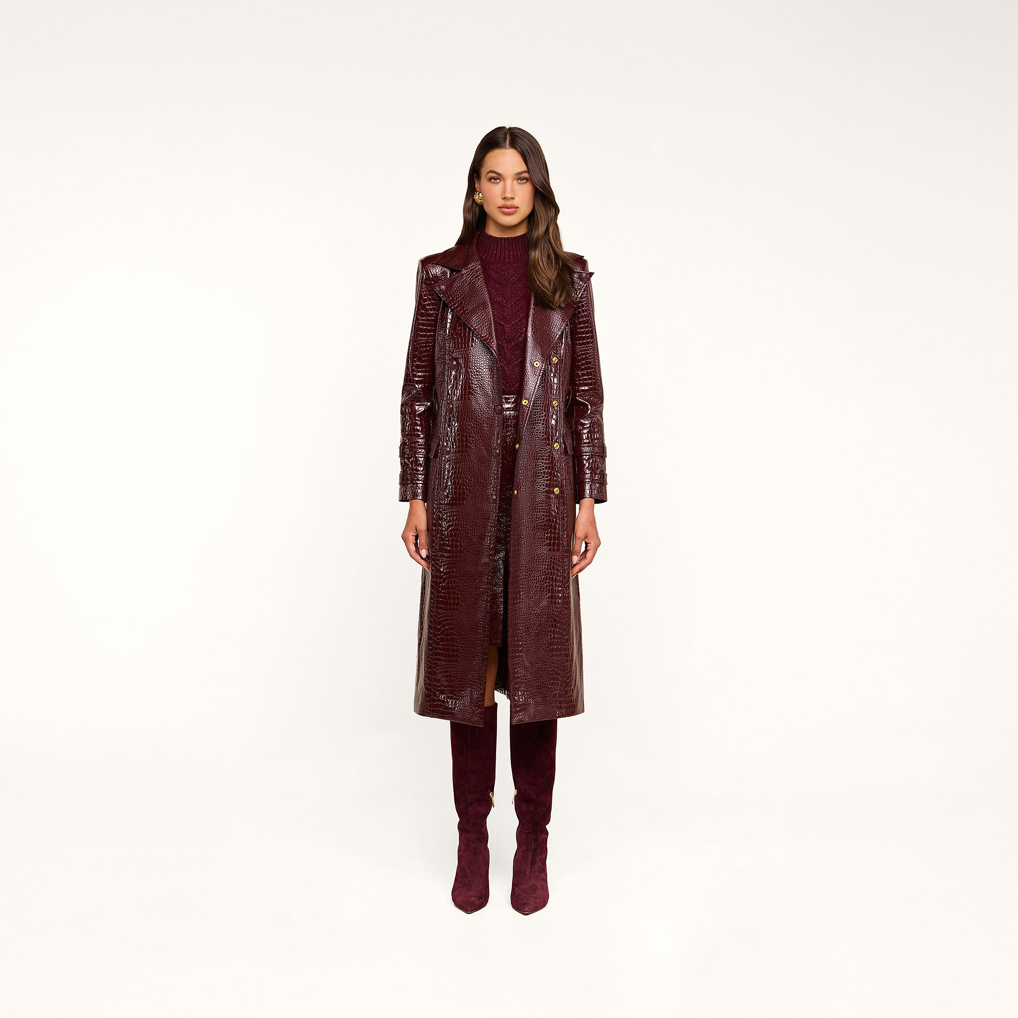 A woman stands facing forward, wearing the Celina Trench Coat—a shiny burgundy vegan leather piece with crocodile embossing—layered over a matching dress and knee-high suede boots, set against a plain beige background for a striking look inspired by Fall 2026 fashion trends.