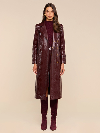 A woman stands facing forward, wearing the Celina Trench Coat—a shiny burgundy vegan leather piece with crocodile embossing—layered over a matching dress and knee-high suede boots, set against a plain beige background for a striking look inspired by Fall 2026 fashion trends.