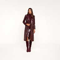 A woman stands wearing the Celina Trench Coat—an elegant, long maroon vegan leather trench featuring subtle crocodile embossing—paired with a matching maroon turtleneck sweater, a metallic silver skirt, and maroon knee-high boots against a plain beige background for an effortlessly chic look inspired by Fall 2026 fashion.