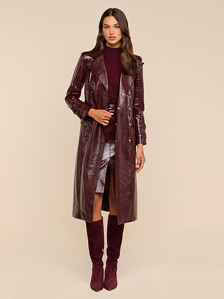 A woman stands wearing the Celina Trench Coat—an elegant, long maroon vegan leather trench featuring subtle crocodile embossing—paired with a matching maroon turtleneck sweater, a metallic silver skirt, and maroon knee-high boots against a plain beige background for an effortlessly chic look inspired by Fall 2026 fashion.
