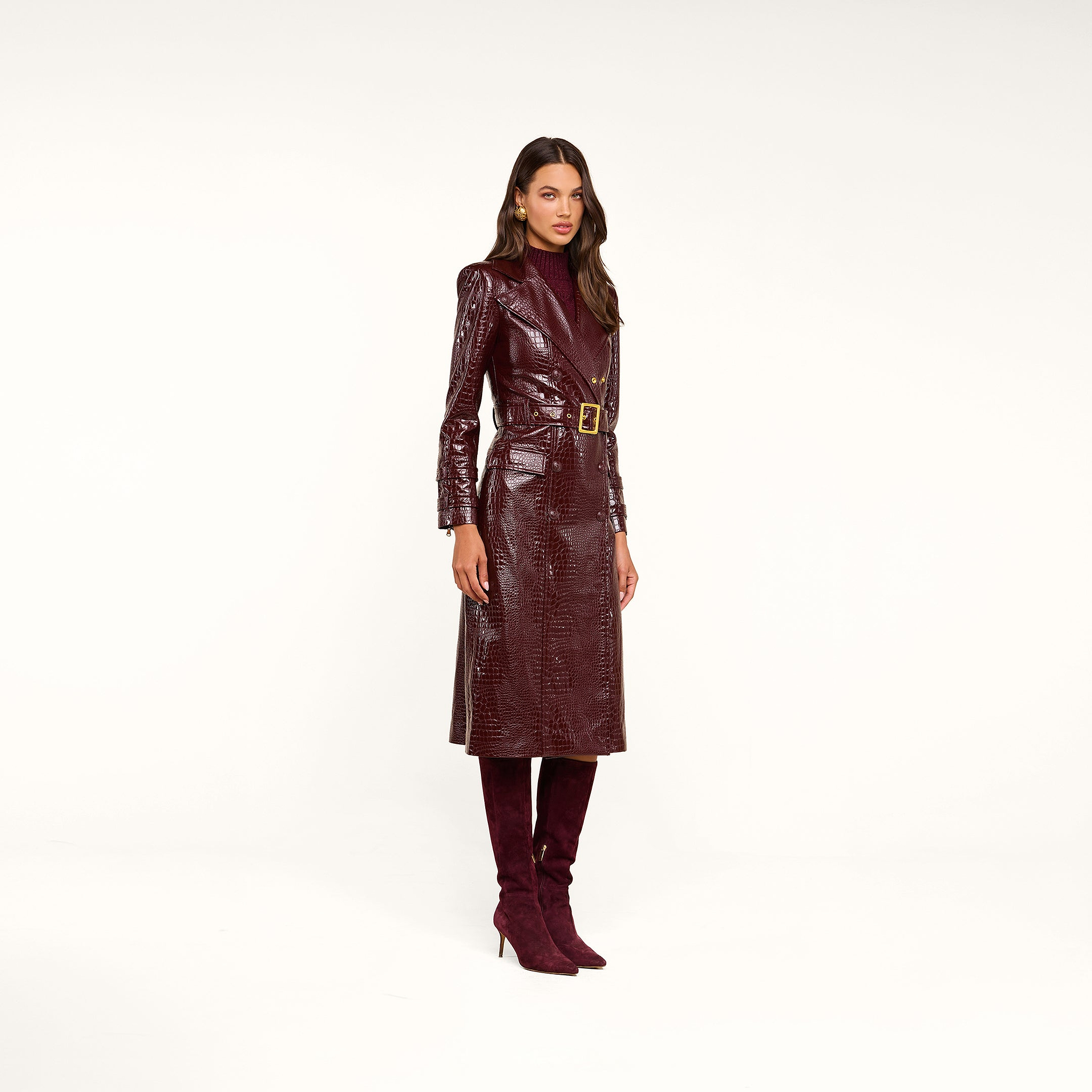 A woman models the Celina Trench Coat—a long, shiny burgundy vegan leather piece with subtle crocodile embossing and a belted waist—styled with a dark turtleneck and matching knee-high heeled boots, standing against a plain beige background for an effortlessly chic, Fall 2026-inspired look.