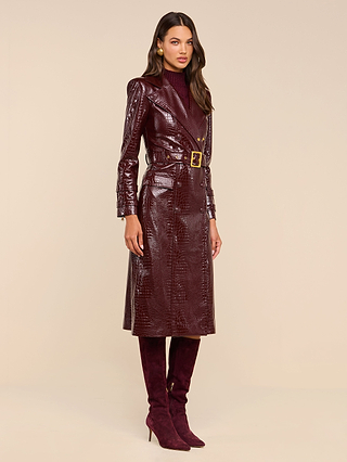 A woman models the Celina Trench Coat—a long, shiny burgundy vegan leather piece with subtle crocodile embossing and a belted waist—styled with a dark turtleneck and matching knee-high heeled boots, standing against a plain beige background for an effortlessly chic, Fall 2026-inspired look.