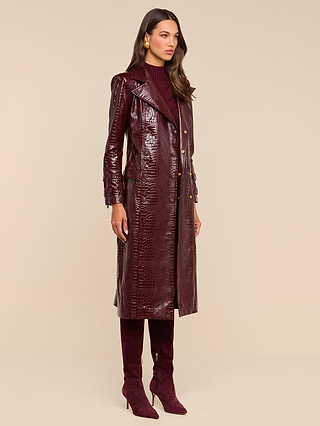 Wearing the Celina Trench Coat—a shiny, burgundy, vegan leather piece with crocodile embossing—paired with matching knee-high heeled boots, a burgundy turtleneck, and gold earrings, this woman stands against a beige background and gazes to the side, embodying a standout look inspired by Fall 2026 fashion trends.