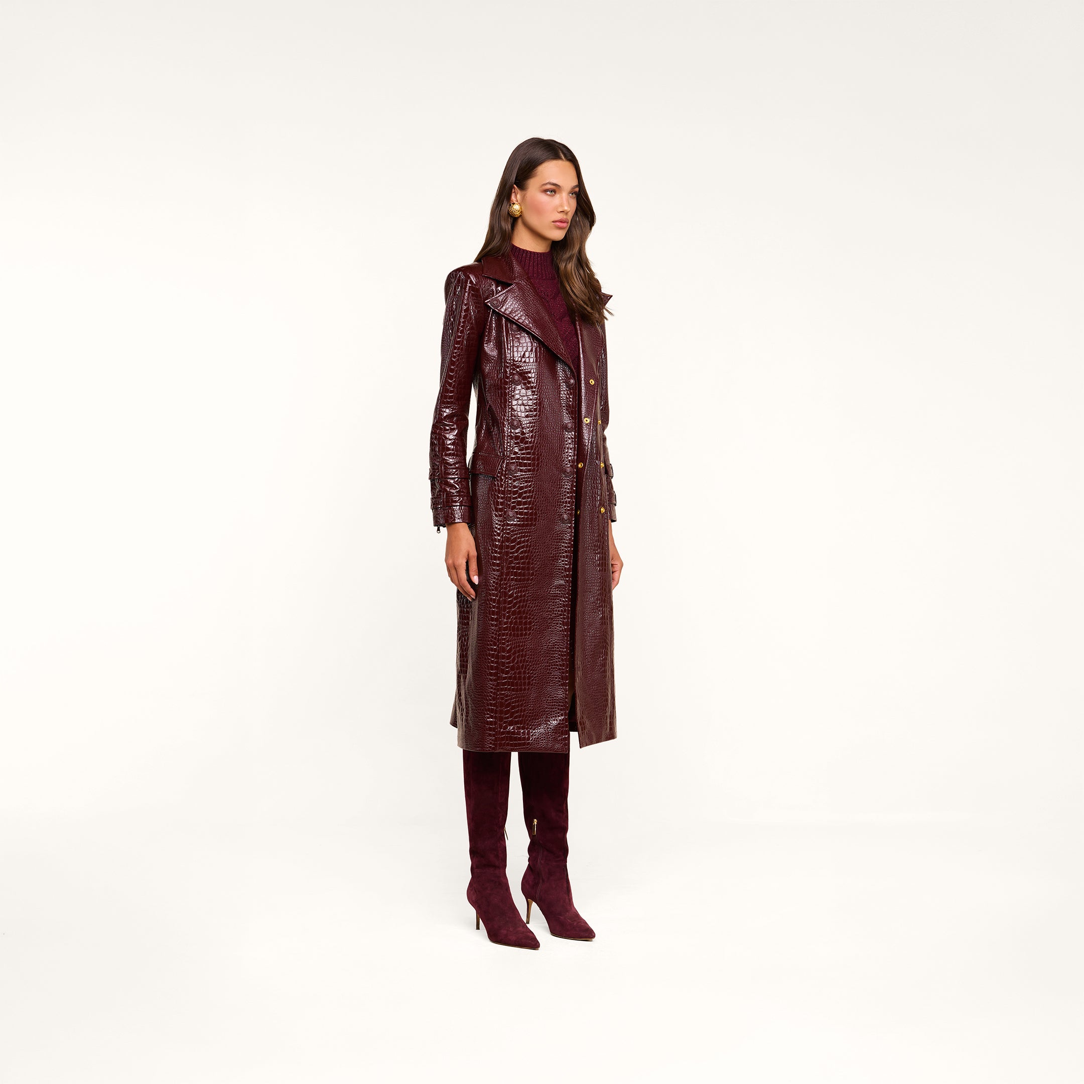 Wearing the Celina Trench Coat—a shiny, burgundy, vegan leather piece with crocodile embossing—paired with matching knee-high heeled boots, a burgundy turtleneck, and gold earrings, this woman stands against a beige background and gazes to the side, embodying a standout look inspired by Fall 2026 fashion trends.