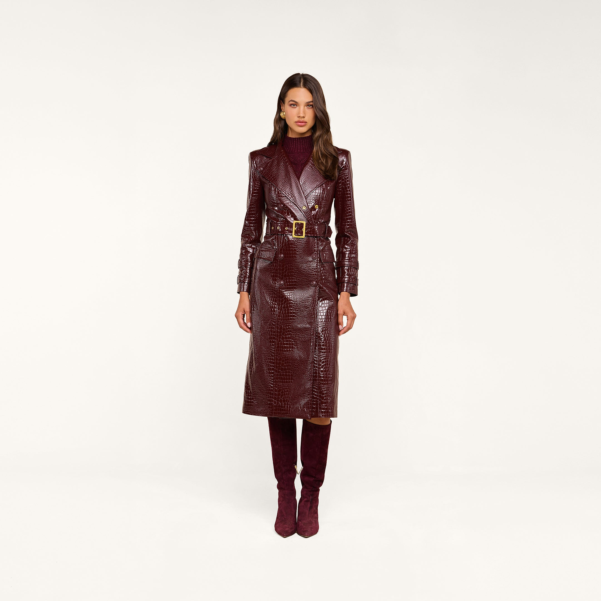 Wearing the Celina Trench Coat in shiny burgundy vegan leather with crocodile embossing and a belted waist, paired with matching knee-high boots and a maroon turtleneck, this woman stands against a plain beige background—a striking look for Fall 2026 fashion.