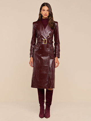 Wearing the Celina Trench Coat in shiny burgundy vegan leather with crocodile embossing and a belted waist, paired with matching knee-high boots and a maroon turtleneck, this woman stands against a plain beige background—a striking look for Fall 2026 fashion.