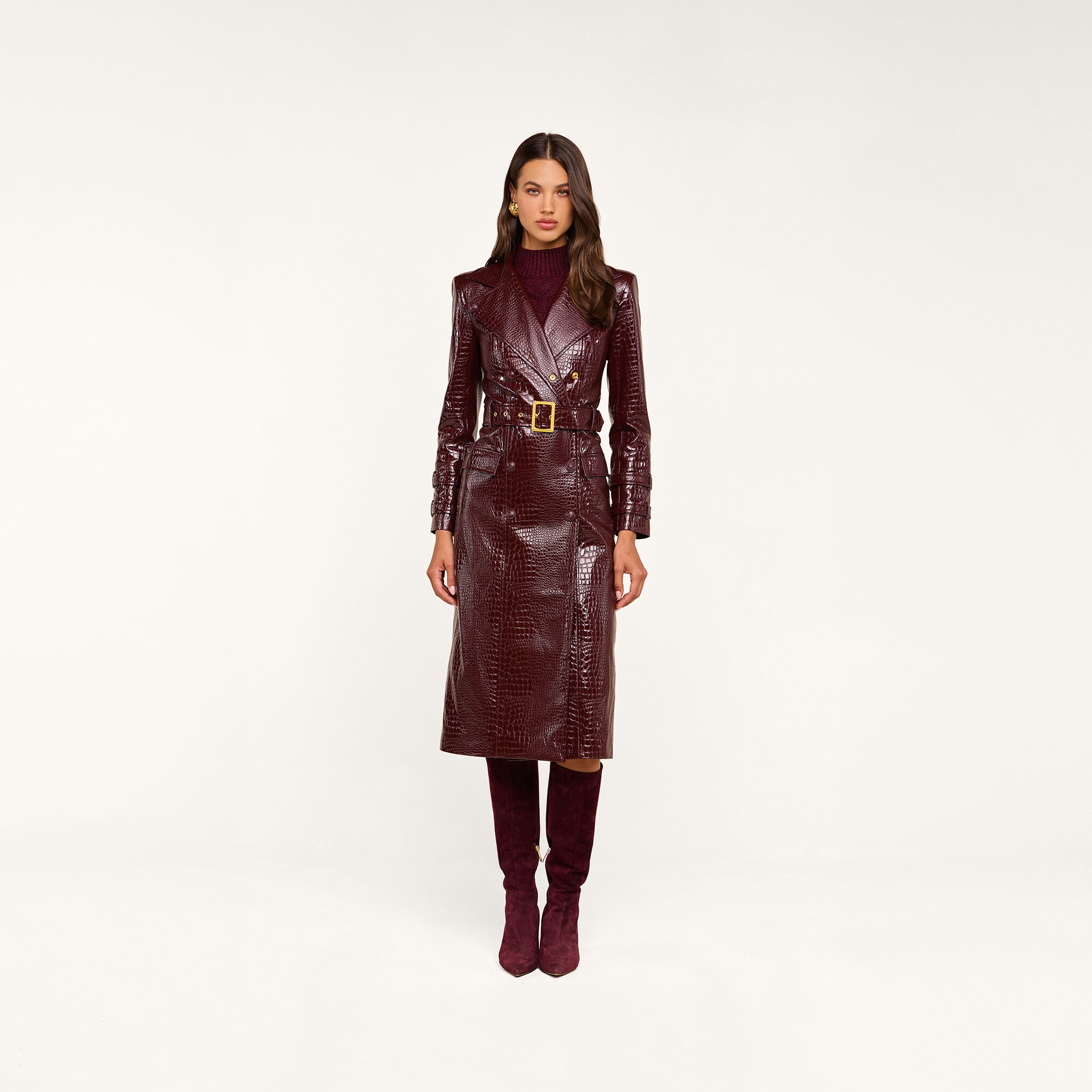 Wearing the Celina Trench Coat in shiny burgundy vegan leather with crocodile embossing and a belted waist, paired with matching knee-high boots and a maroon turtleneck, this woman stands against a plain beige background—a striking look for Fall 2026 fashion.