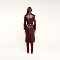 A woman stands facing away, wearing the Celina Trench Coat—a long, glossy, dark red vegan leather trench coat with crocodile embossing and a belted waist—paired with matching tall boots. Set against a light beige background, the look captures the bold sophistication of Fall 2026 fashion.