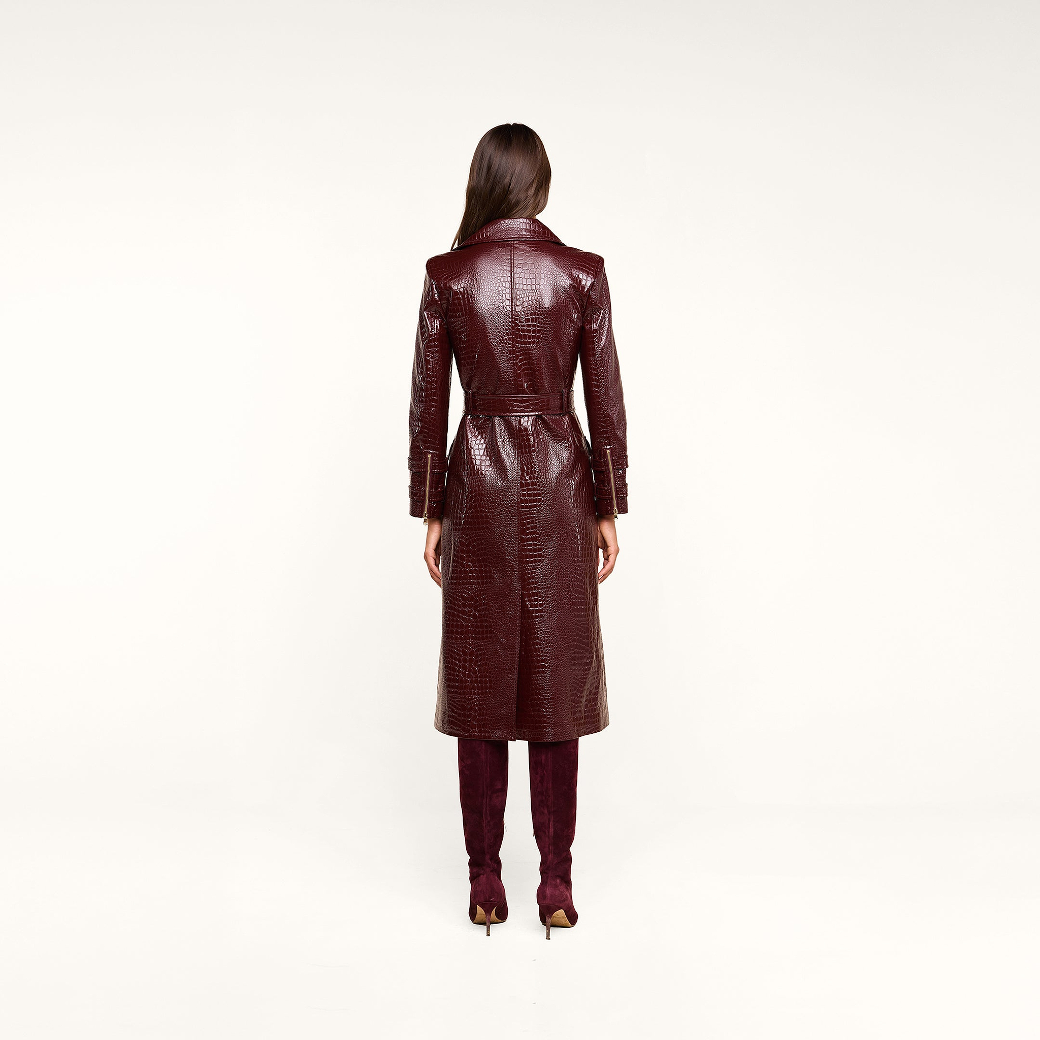 A woman stands facing away, wearing the Celina Trench Coat—a long, glossy, dark red vegan leather trench coat with crocodile embossing and a belted waist—paired with matching tall boots. Set against a light beige background, the look captures the bold sophistication of Fall 2026 fashion.