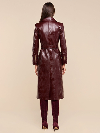 A woman stands facing away, wearing the Celina Trench Coat—a long, glossy, dark red vegan leather trench coat with crocodile embossing and a belted waist—paired with matching tall boots. Set against a light beige background, the look captures the bold sophistication of Fall 2026 fashion.