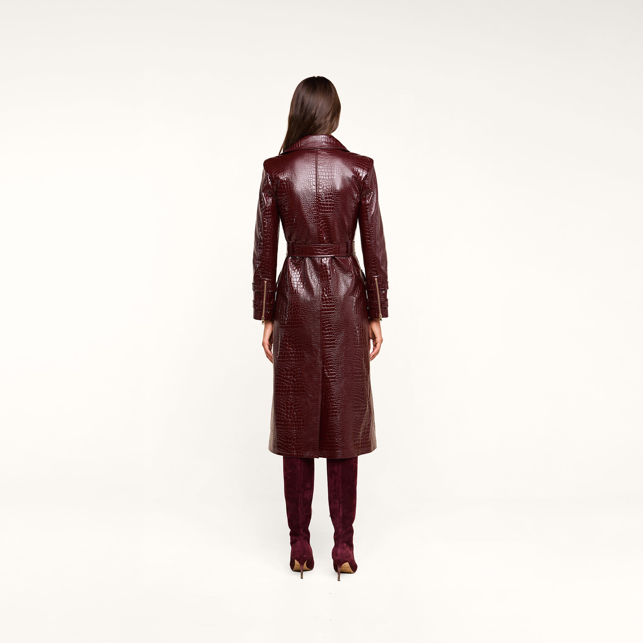 A woman stands facing away, wearing the Celina Trench Coat—a long, glossy, dark red vegan leather trench coat with crocodile embossing and a belted waist—paired with matching tall boots. Set against a light beige background, the look captures the bold sophistication of Fall 2026 fashion.