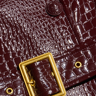 Close-up of a dark brown, crocodile-patterned leather bag with a shiny finish, featuring a rectangular gold buckle and metal eyelets—a perfect accessory to complement the Celina Trench Coat for Fall 2026 fashion and vegan leather trench looks with its elegant crocodile embossing.