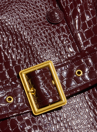 Close-up of a dark brown, crocodile-patterned leather bag with a shiny finish, featuring a rectangular gold buckle and metal eyelets—a perfect accessory to complement the Celina Trench Coat for Fall 2026 fashion and vegan leather trench looks with its elegant crocodile embossing.