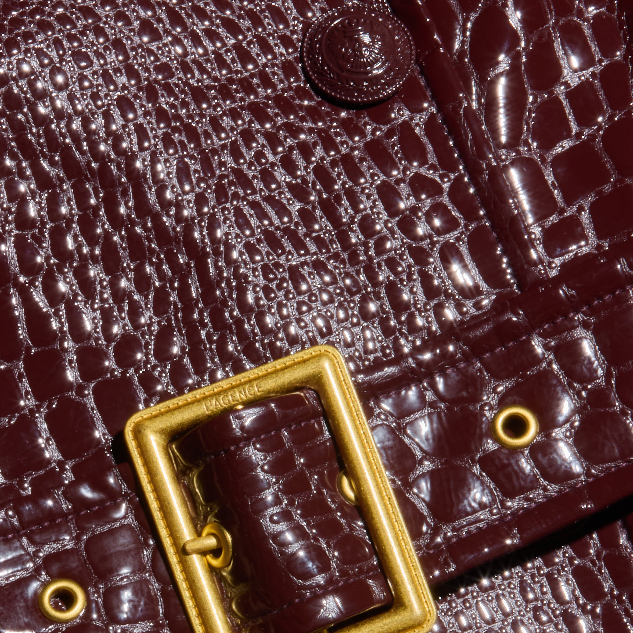 Close-up of a dark brown, crocodile-patterned leather bag with a shiny finish, featuring a rectangular gold buckle and metal eyelets—a perfect accessory to complement the Celina Trench Coat for Fall 2026 fashion and vegan leather trench looks with its elegant crocodile embossing.