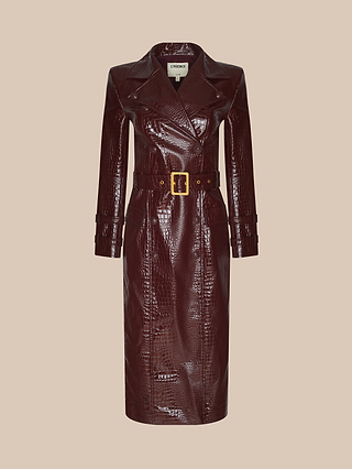 The Celina Trench Coat, crafted from long, dark burgundy vegan leather with crocodile embossing, showcases wide lapels, a coordinating belt with a gold buckle, and buttoned cuffs. Displayed against a beige background, it makes a bold statement for Fall 2026 fashion.