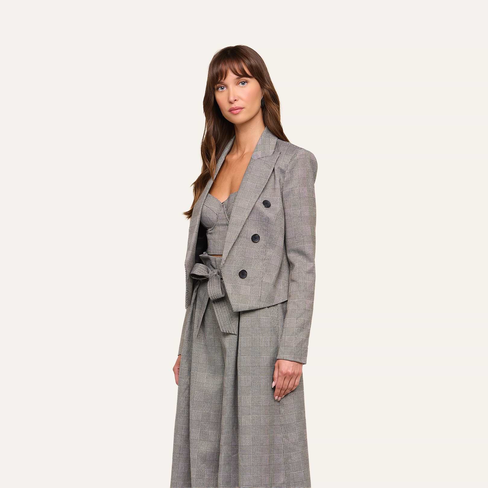 A woman wearing the Lila Tweed Blazer—a gray plaid jacket featuring a belted waist and three-button closure—stands against a plain light background.