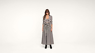 A woman wearing sunglasses, a long plaid dress, and a matching blazer stands against a plain white background. She has long hair and is wearing black ankle boots.